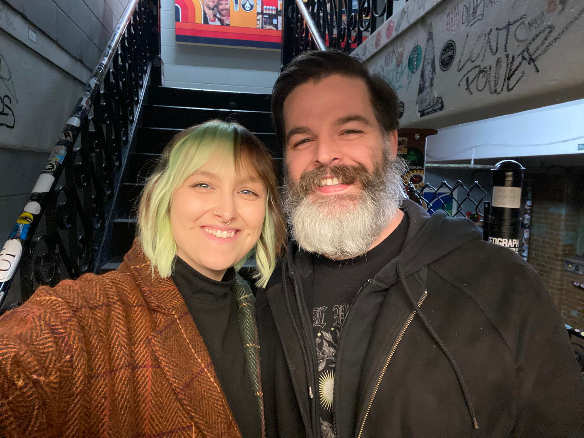 two humans standing at the bottom of a grimy staircase. the woman on the left has green and brown hair, the man on the right has dark hair and a greying beard.