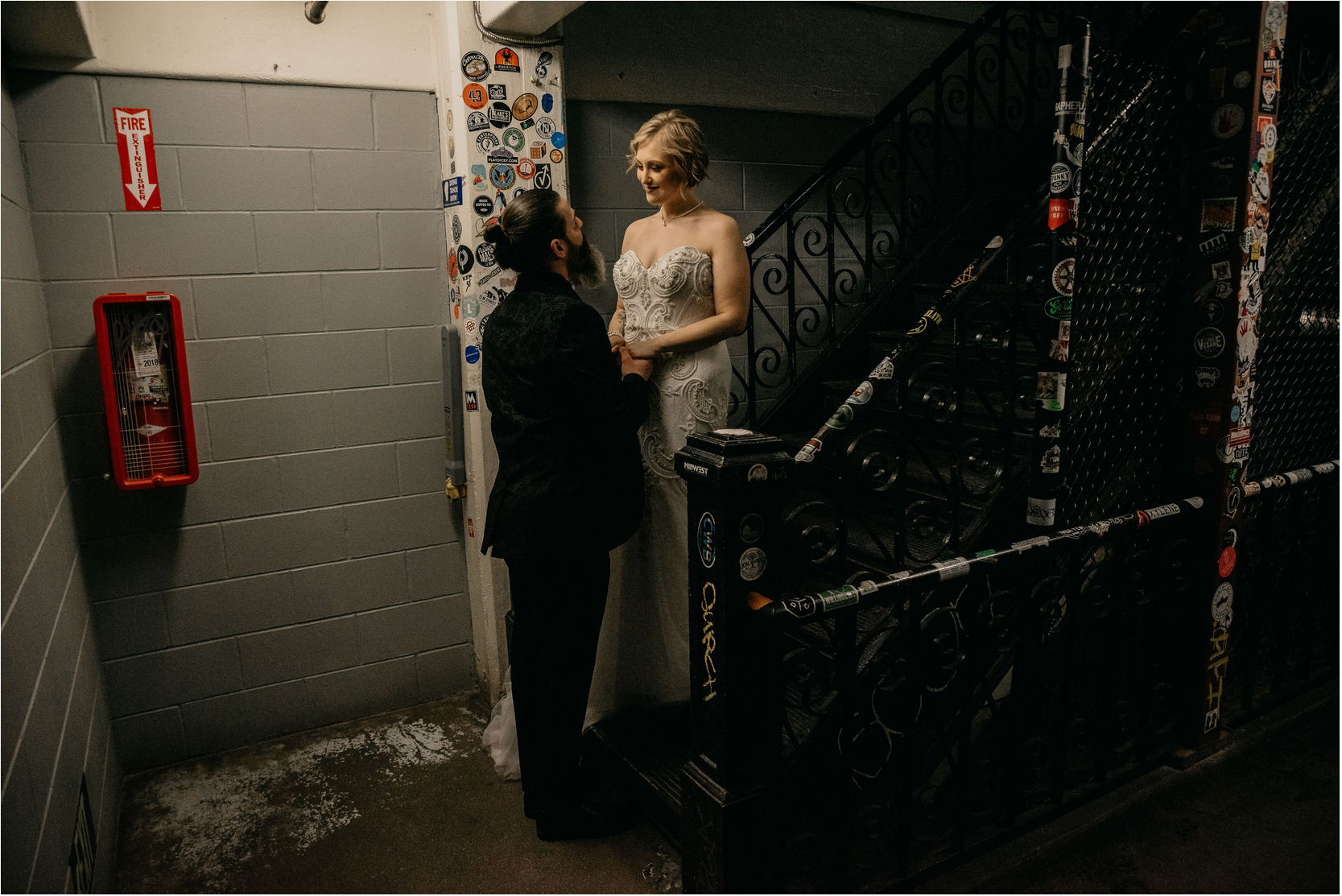 two humans standing at the bottom of a grimy staircase. the woman on the left has green and brown hair, the man on the right has dark hair and a greying beard.