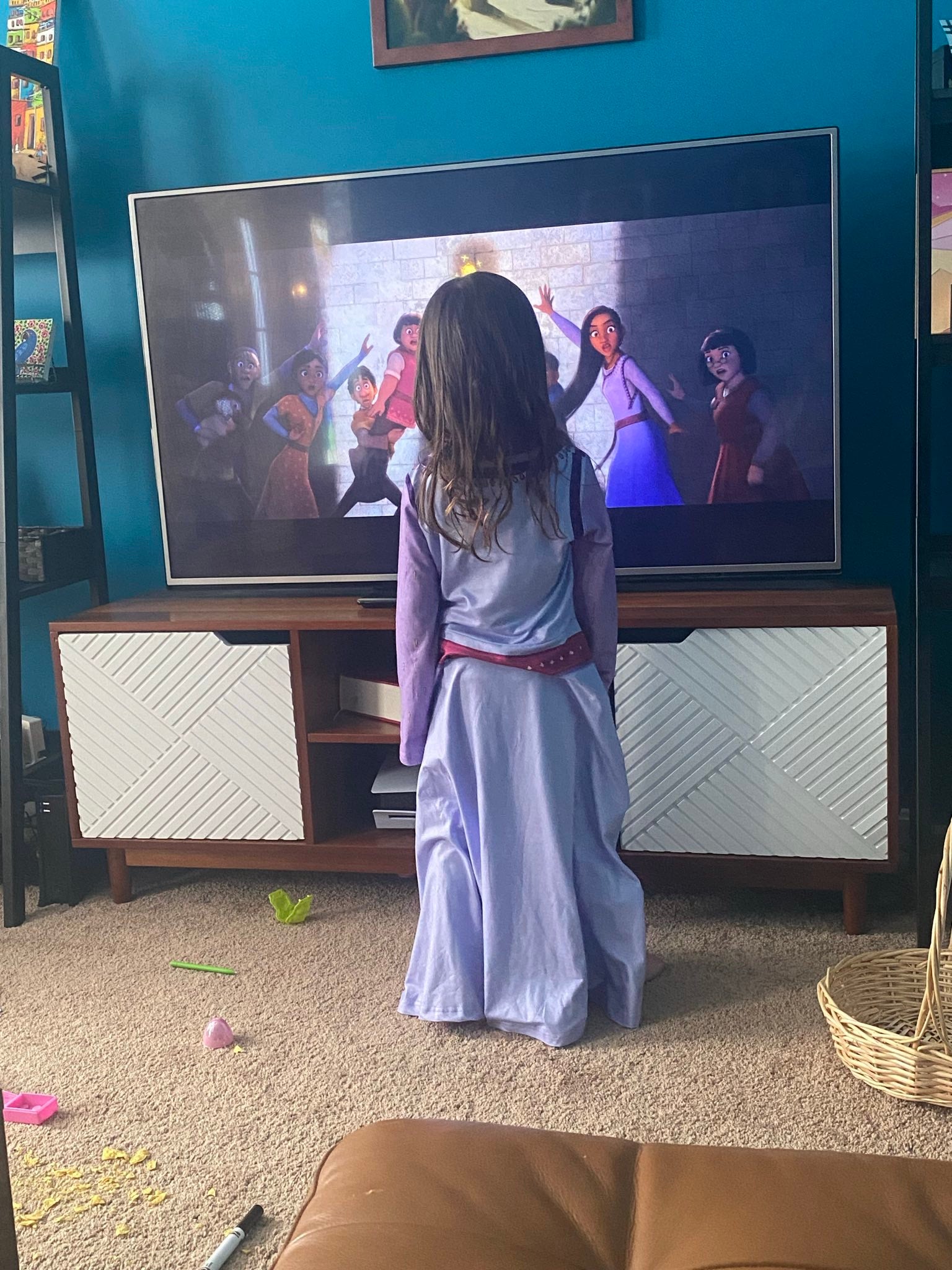 4 year old girl with brown hair faces a tv playing Wish wearing an oversized Asha’s purple dress